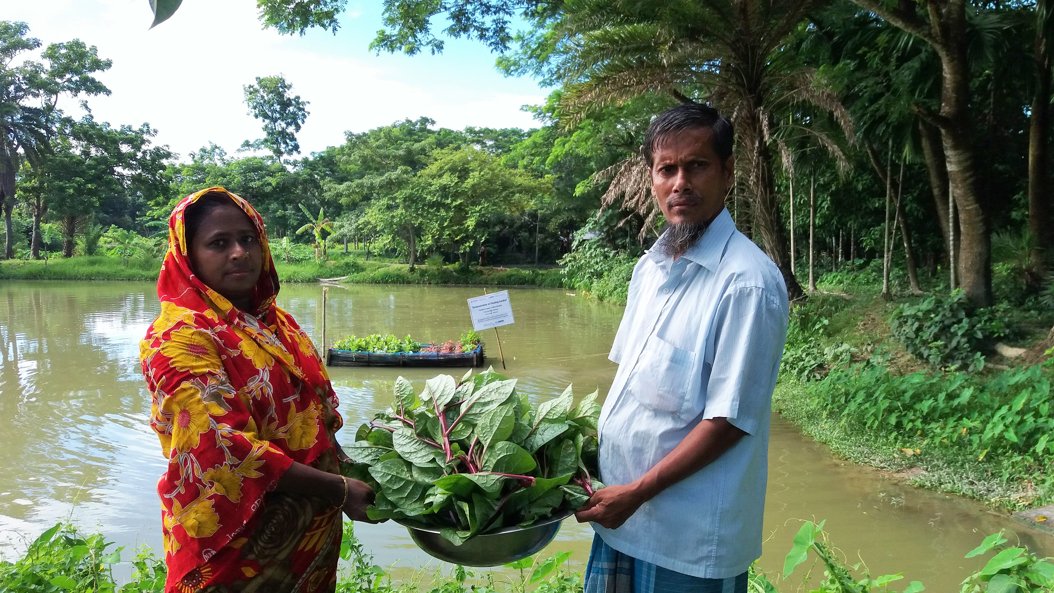 Man and woman hold basket of leafy green vegetables in front of pond with floating garden in Bangladesh