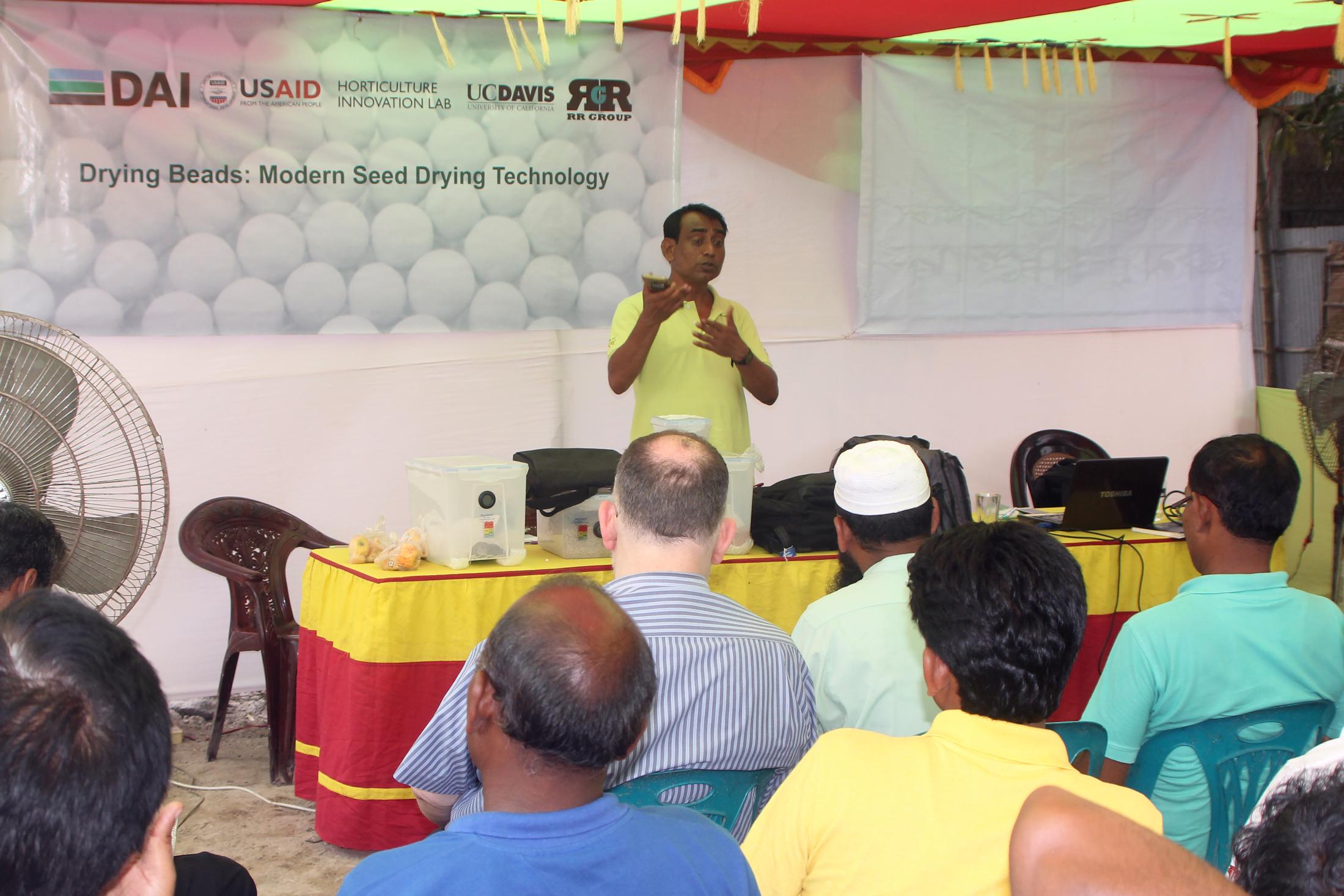  Man holds up drying beads to teach crowd with banner DAI USAID logos in background.