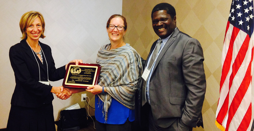 Two women shake hands, holding a plaque, man stands next to them, all smiling