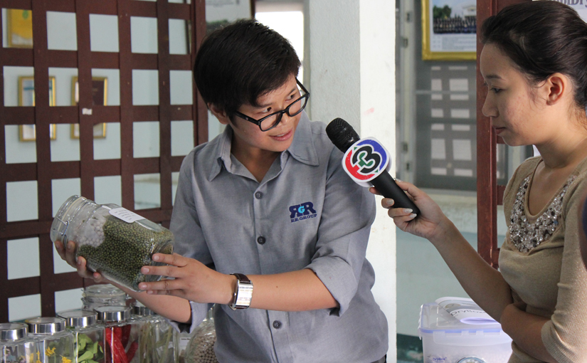 Woman holds jar of drying beads and seeds, speaking into TV microphone held by a reporter