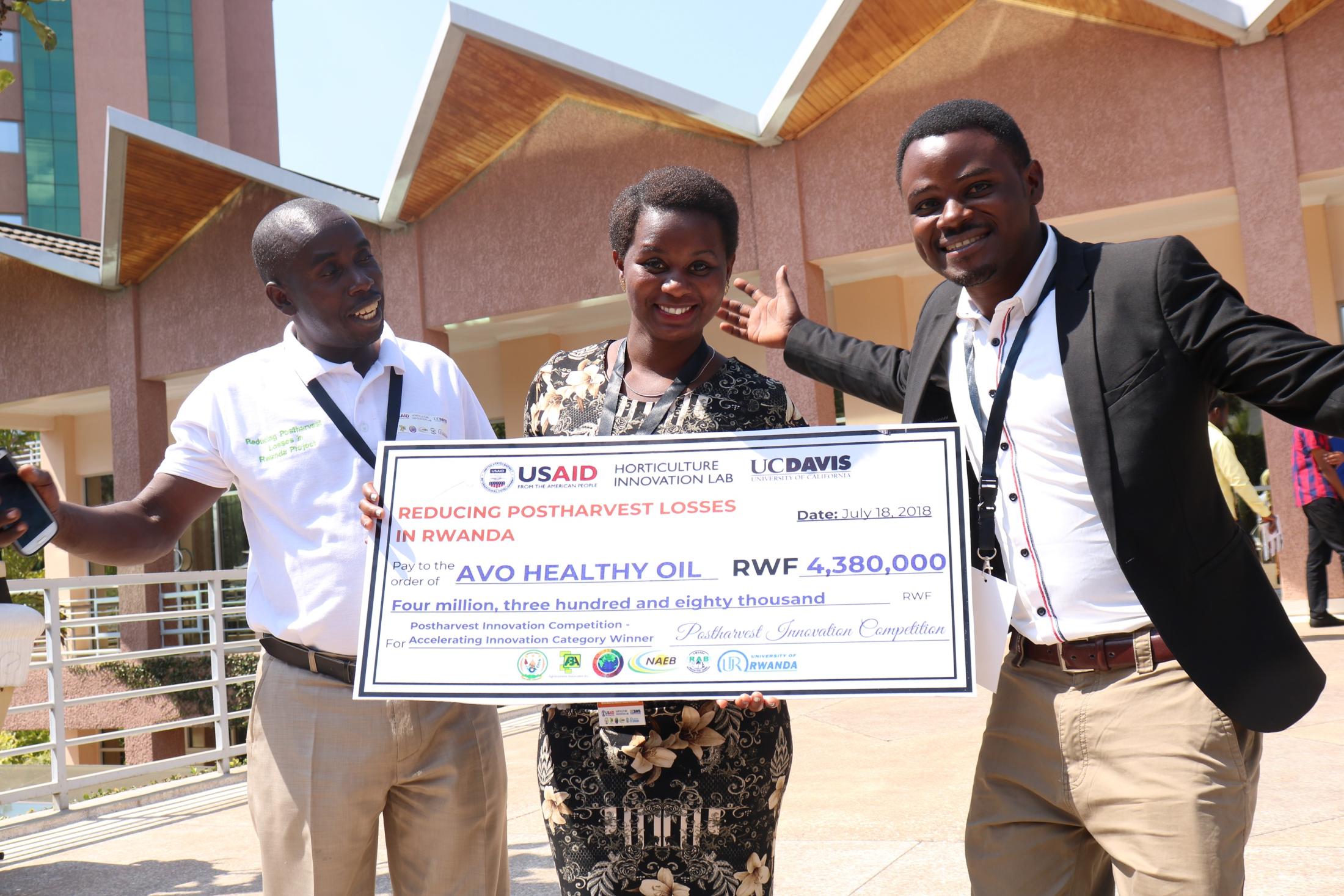 Niyidukunda Mugeni Euphrosine of Avo Healthy Oil Company grins and holds a giant check showing her award money from the competition, next to two other competitors.