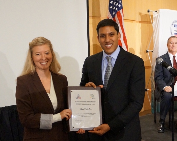 Elana accepts award plaque from USAID leader, American flag in background