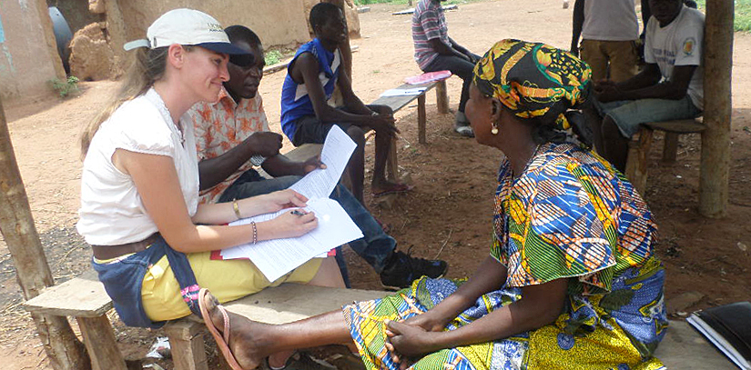 Smiling researcher takes notes while farmer speaks, seated in shade