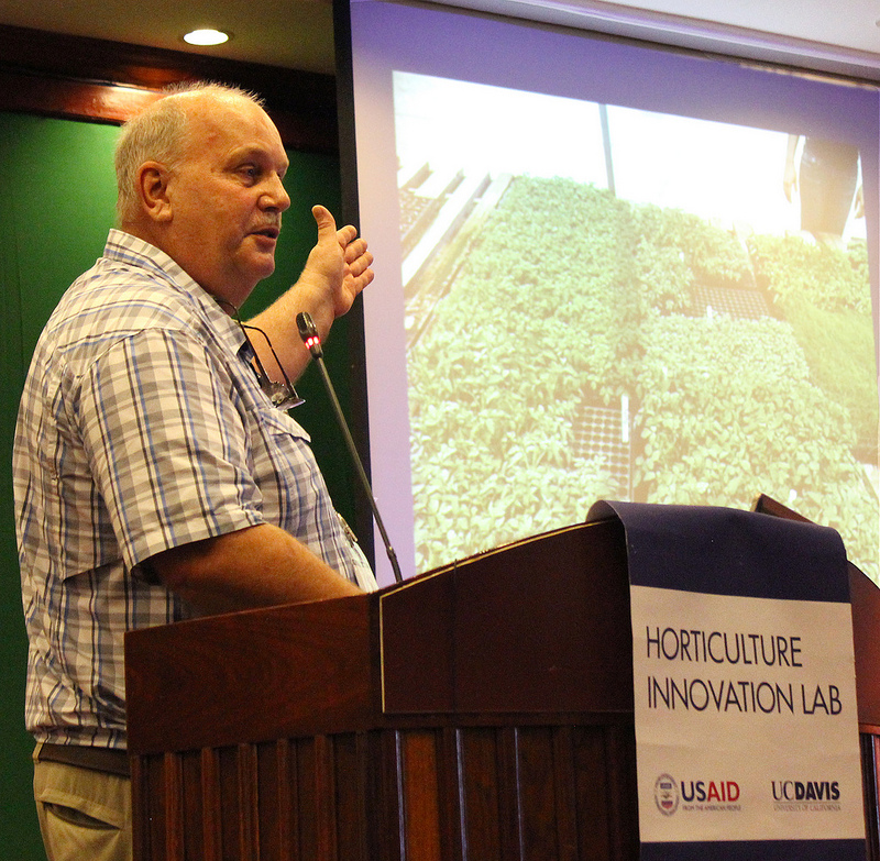 Man speaking at podium, gesturing to powerpoint slide with greenhouse nursery photo