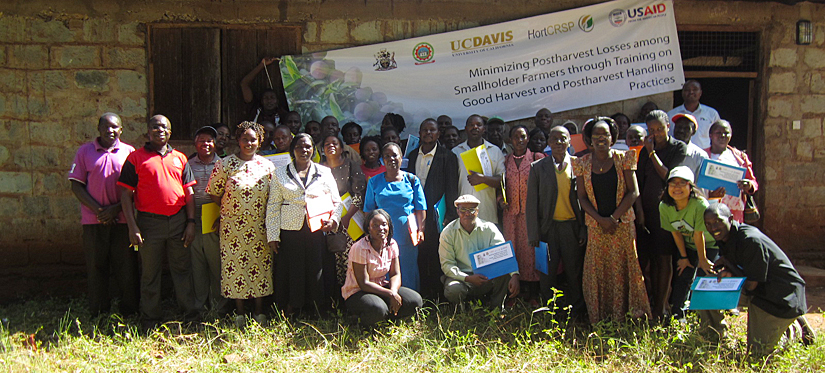 smiling group of farmers and researchers outdoors, in front of school house with usaid logo banner