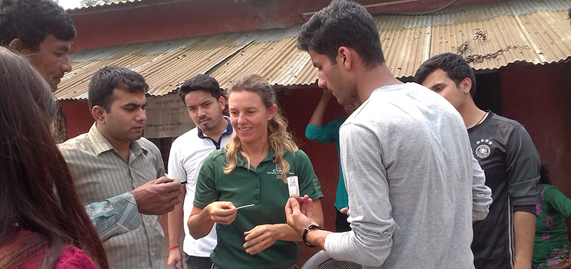 Grad student woman looking at paper test indicator offered by a Nepalese man, with group looking on at farm