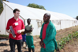 Three men stand in front of a greenhouse in a field