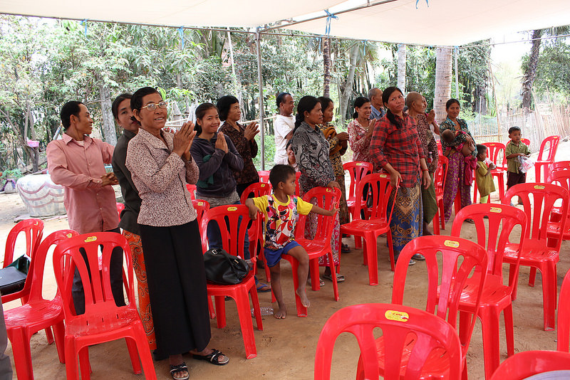 Group of approx. 20 people under a tent stand around red chairs