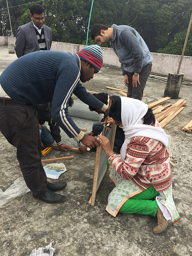 Four people wield tools and build with pieces of wood, two men look on