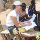 Smiling researcher takes notes while farmer speaks, seated in shade